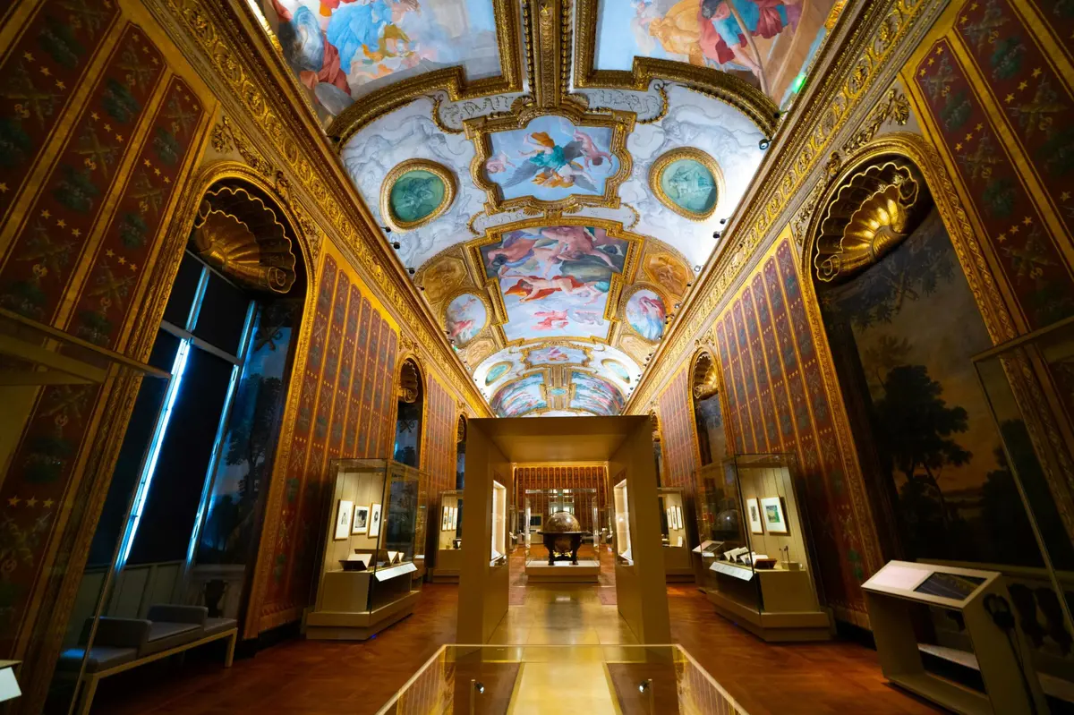 Ornate museum gallery with intricate ceiling paintings, gold accents, and illuminated display cases showing artwork and artifacts.