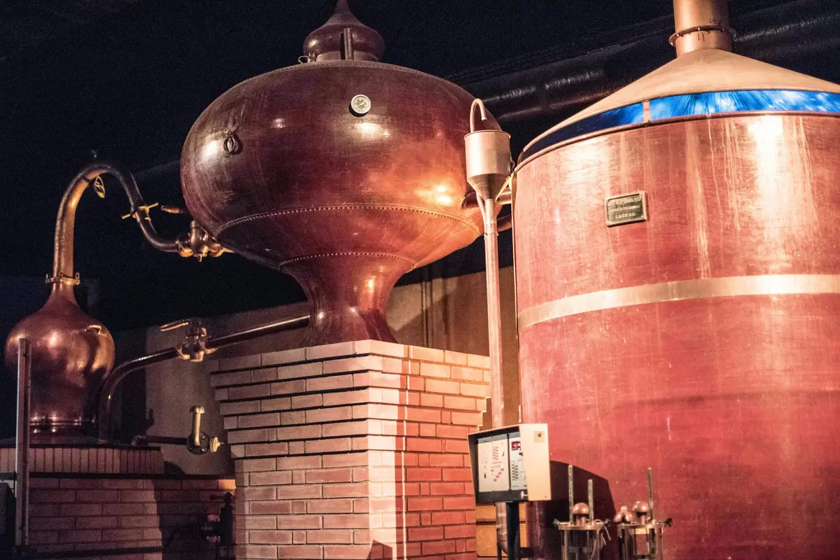 Grandes cuves de distillation en cuivre avec des bases en briques à l'intérieur d'une distillerie, avec des tuyaux et un panneau de contrôle.