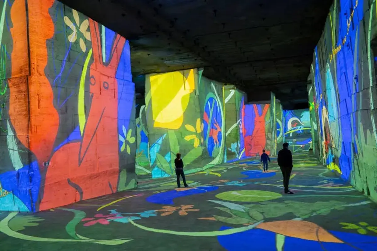 Des personnes traversent un grand espace intérieur avec des projections colorées d'œuvres d'art abstraites et inspirées de la nature sur les murs et le sol.