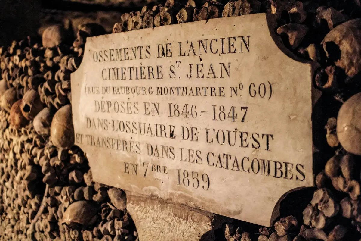 Une plaque située dans les Catacombes de Paris, entourée d'ossements empilés, sur laquelle figure un texte en français fournissant des informations historiques sur les sépultures.