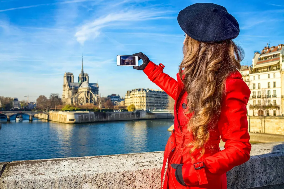 Personne vêtue d'un manteau rouge et d'un béret noir prenant une photo à l'aide d'un smartphone, capturant un point de repère au bord de la rivière sous un ciel bleu.