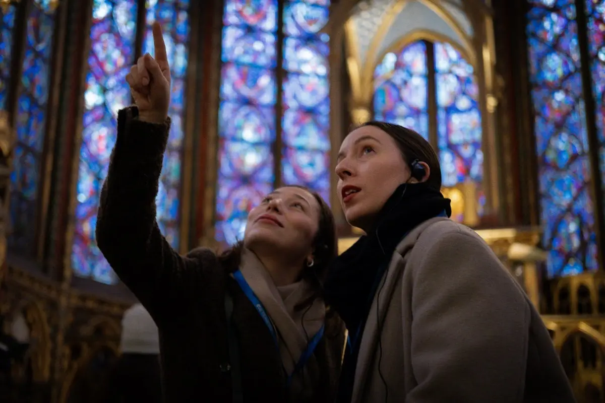 Deux femmes avec des écouteurs regardent vers le haut devant des vitraux colorés à l'intérieur d'une cathédrale.