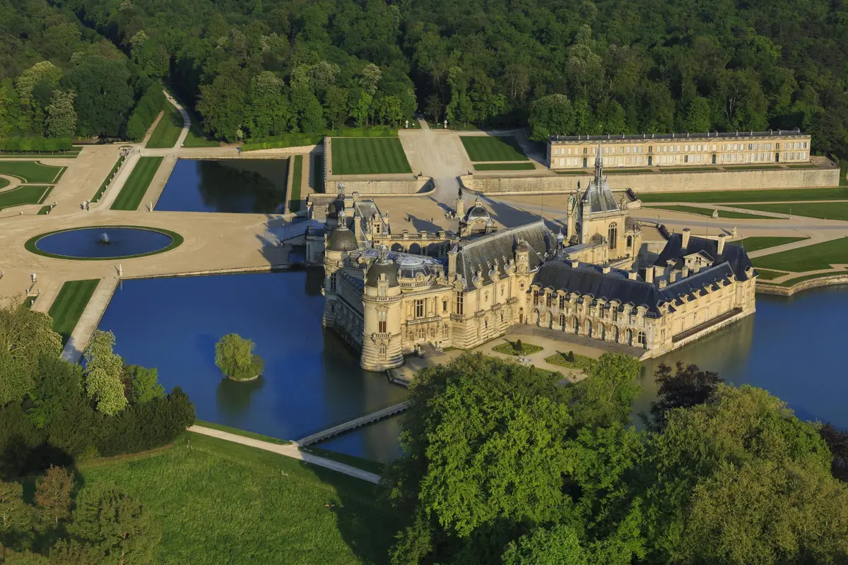 Vue aérienne d'un grand château historique entouré de douves, de jardins luxuriants et d'une forêt dense.