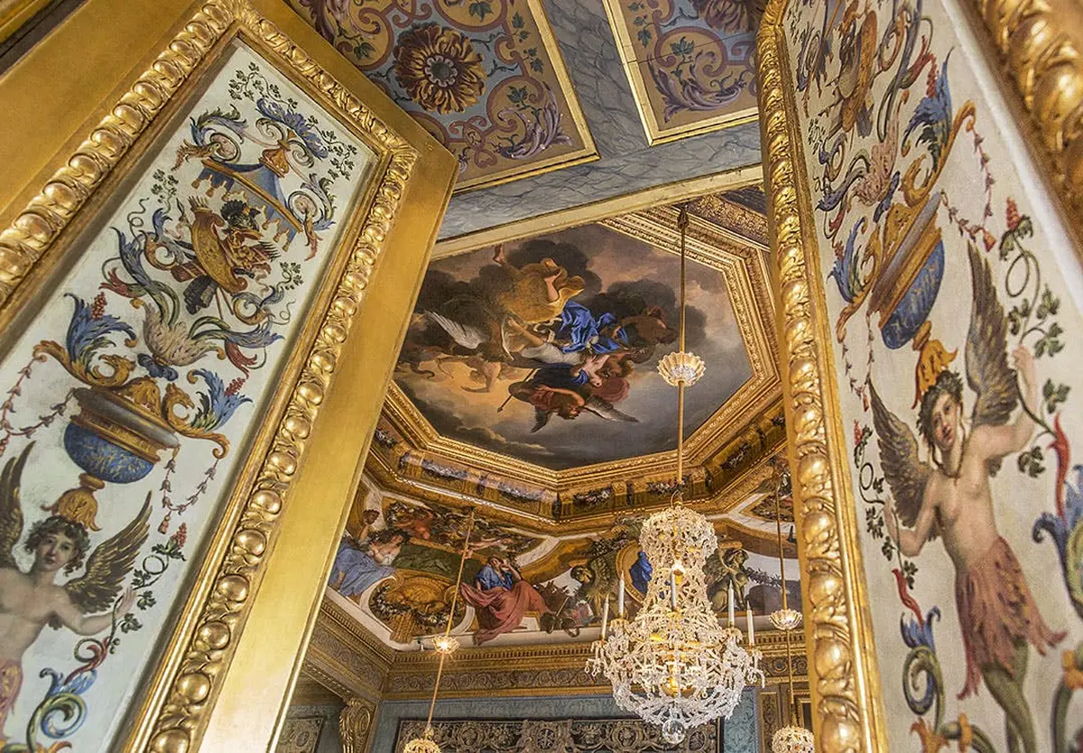 Ornate room with a painted ceiling depicting a mythological scene, golden-framed walls, and a crystal chandelier.