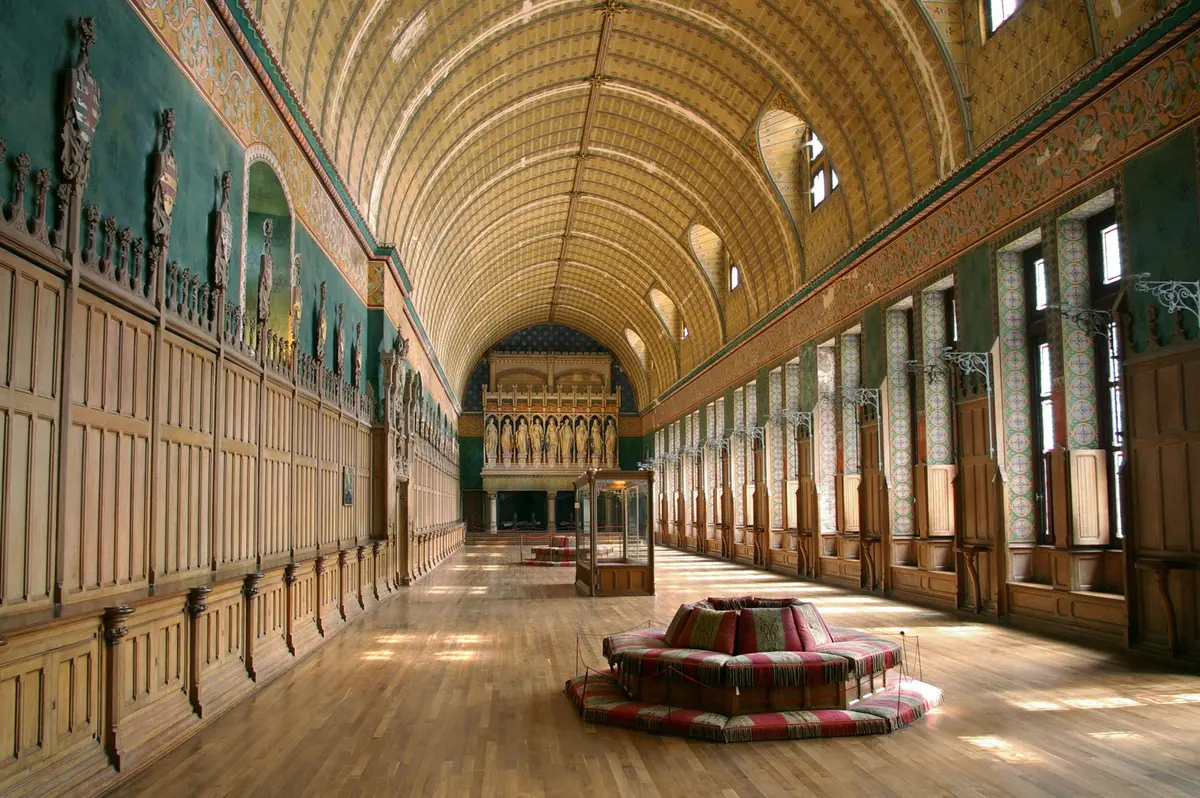 Une grande salle avec un plafond voûté, des décorations murales ornées, un plancher en bois, de hautes fenêtres cintrées et un siège central rembourré.