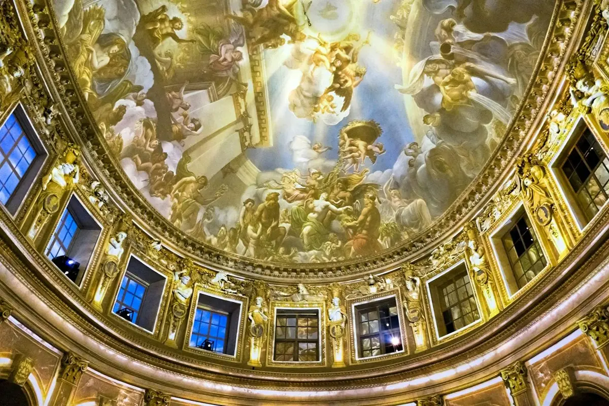 Un plafond en dôme orné de fresques détaillées de personnages et d'un motif architectural doré, entouré de grandes fenêtres.