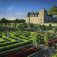 Des jardins manucurés avec des parterres de fleurs et des haies colorées devant un château historique en pierre sous un ciel bleu clair. DR