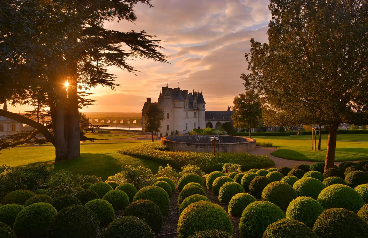Jardin entretenu avec des haies rondes devant un château historique au coucher du soleil, avec de grands arbres et un ciel spectaculaire en arrière-plan.