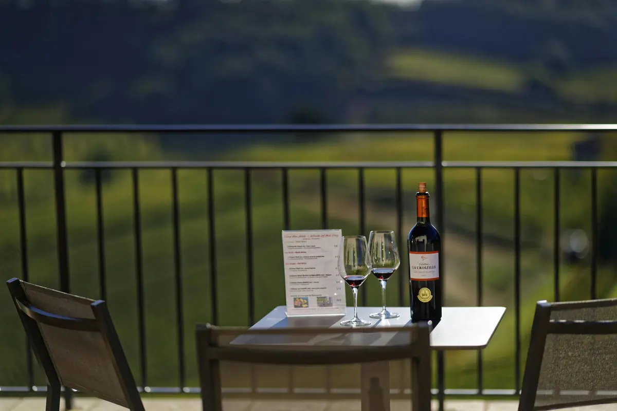 Table avec une bouteille de vin, deux verres de vin rouge et un menu donnant sur un balcon avec des collines vertes floues en arrière-plan.
