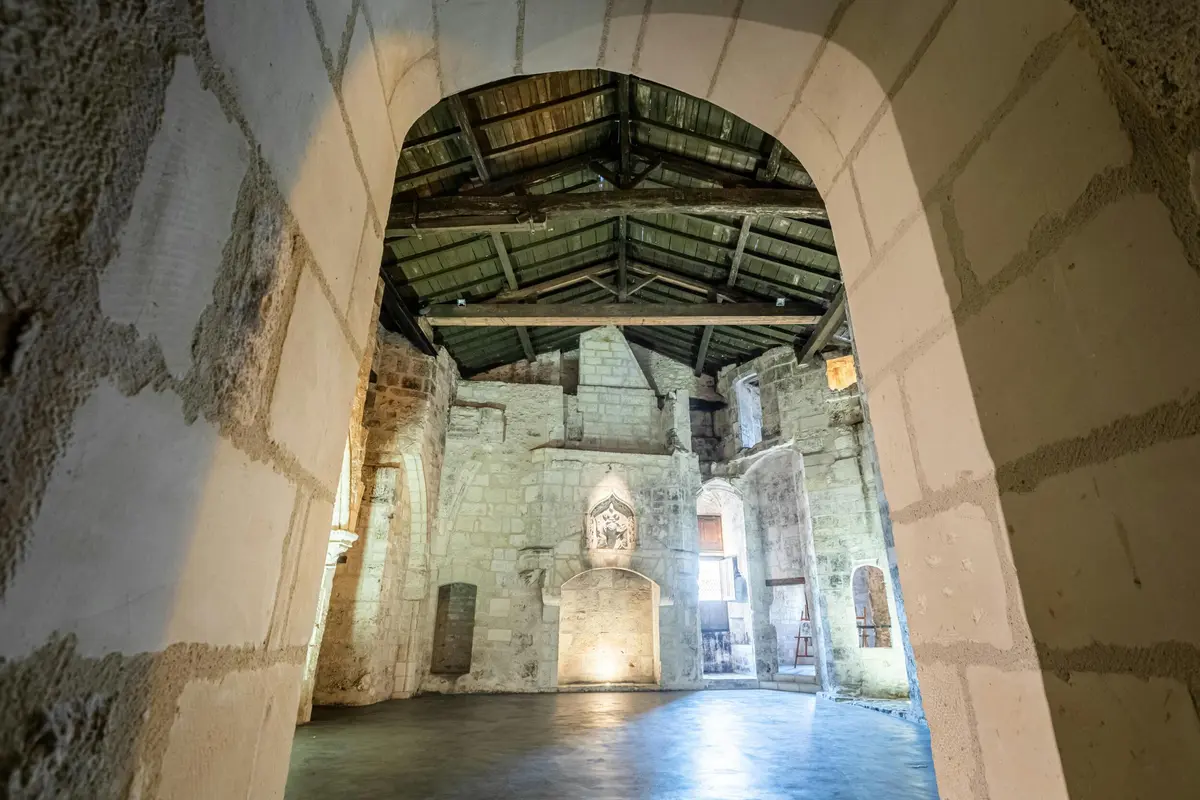 Intérieur d'un bâtiment historique en pierre avec un plafond en poutres de bois, des portes cintrées et un sol lisse, vu à travers une grande arche.
