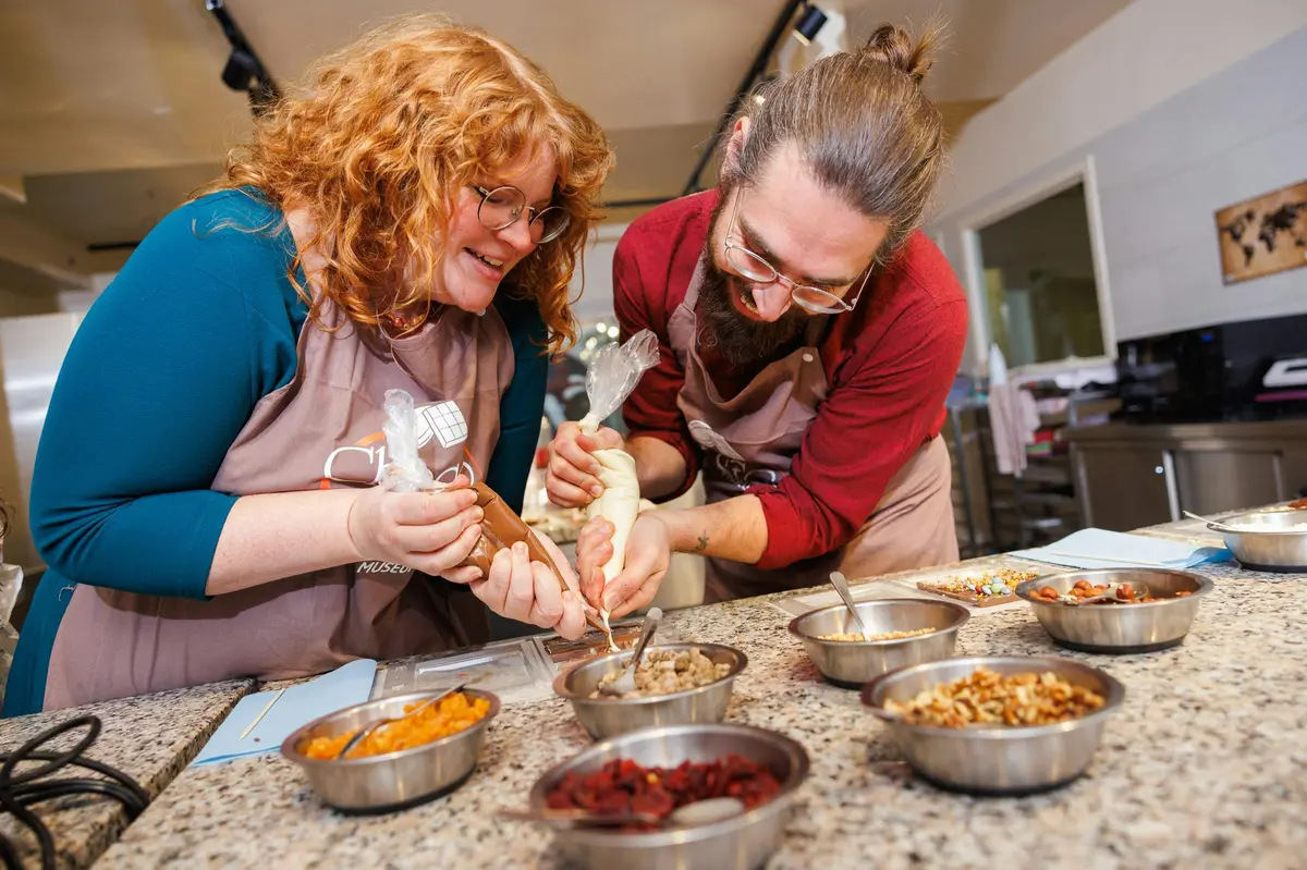 Deux personnes souriant et décorant des aliments avec des poches à douille dans une cuisine, entourées de bols d'ingrédients.