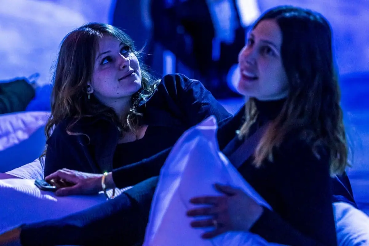 Deux femmes dans une pièce faiblement éclairée par une lumière bleue, l'une tenant un oreiller, toutes deux semblant détendues et engagées.