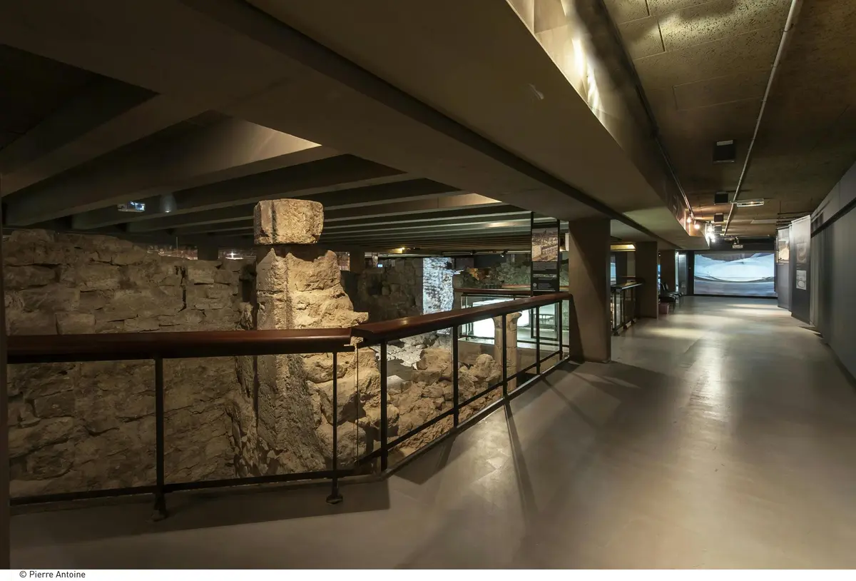 Musée souterrain présentant d'anciennes ruines en pierre, des rampes métalliques et des panneaux d'information sous un plafond en béton.