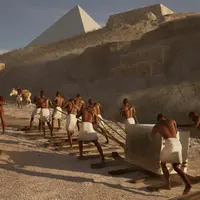 Des personnes vêtues de vêtements blancs tirent un gros bloc de pierre à l'aide de cordes sur des planches de bois près des pyramides. &copy; TDB