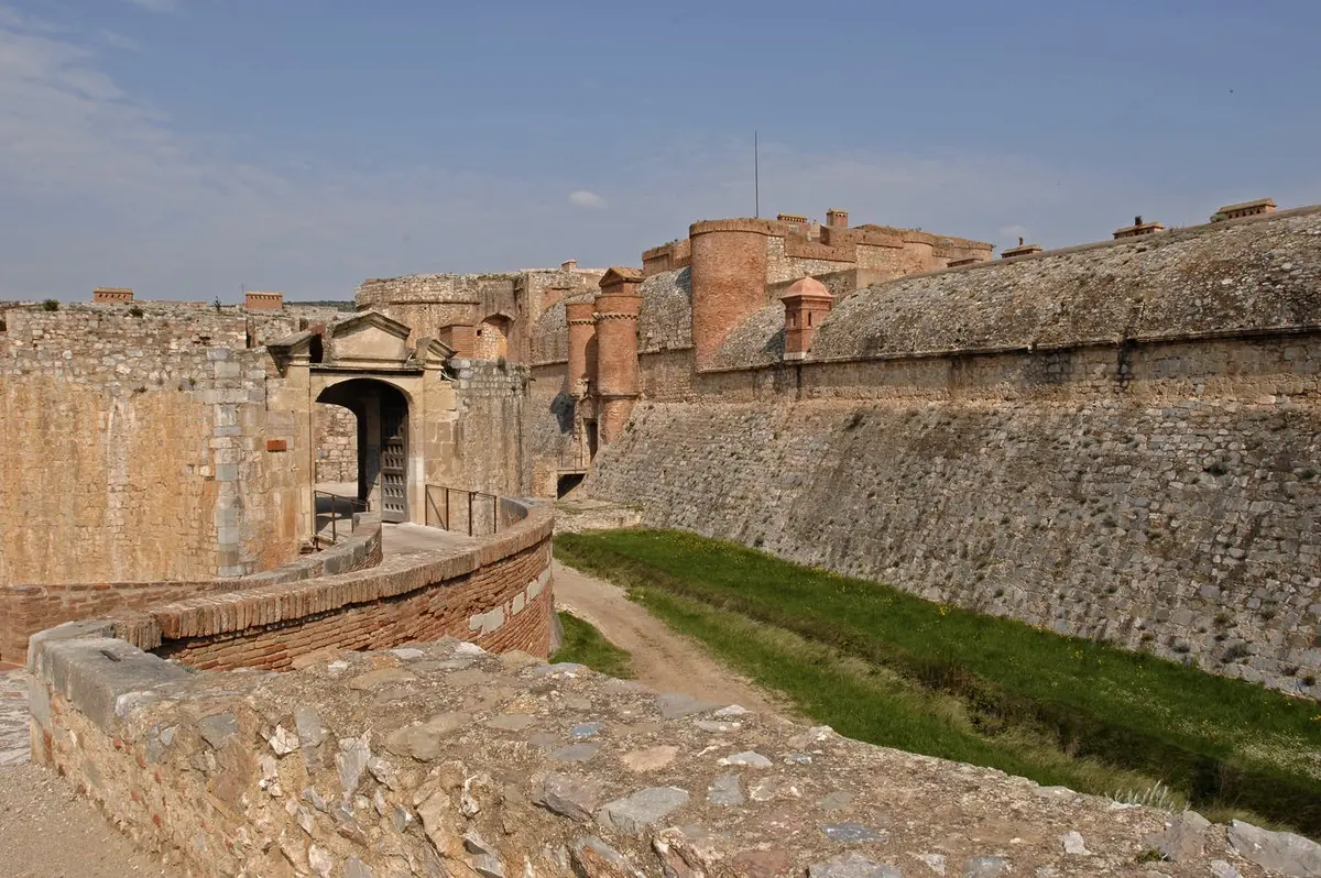 Forteresse historique avec des murs en pierre, une tour de garde, des douves herbeuses et une porte d'entrée sous un ciel bleu clair.