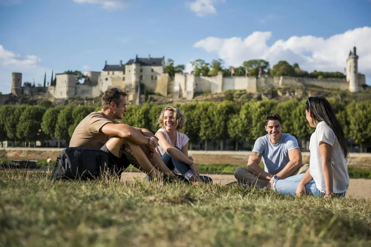 Quatre personnes sont assises et discutent sur l'herbe avec un bâtiment historique et des arbres en arrière-plan sous un ciel bleu avec quelques nuages.