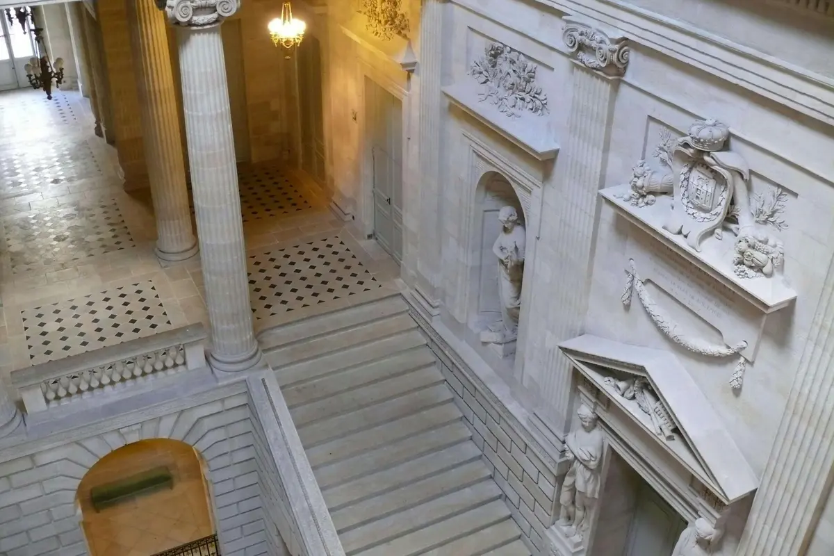 Un escalier en pierre richement orné, situé dans un intérieur somptueux agrémenté de colonnes, de statues dans des niches et de sculptures murales minutieuses, éclairé par un lustre.
