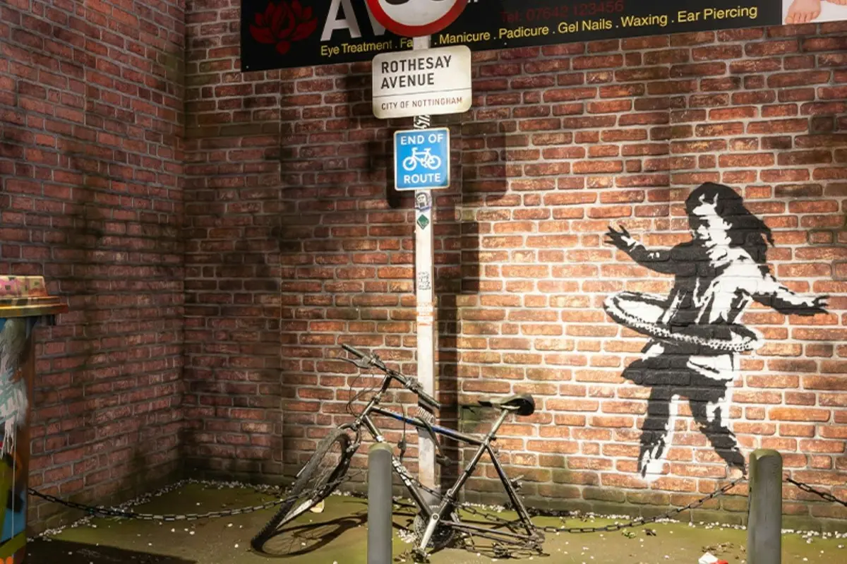 Un vélo est appuyé contre un poteau sur lequel sont accrochés des panneaux, devant un mur de briques orné d'un graffiti représentant une silhouette faisant du hula-hoop.