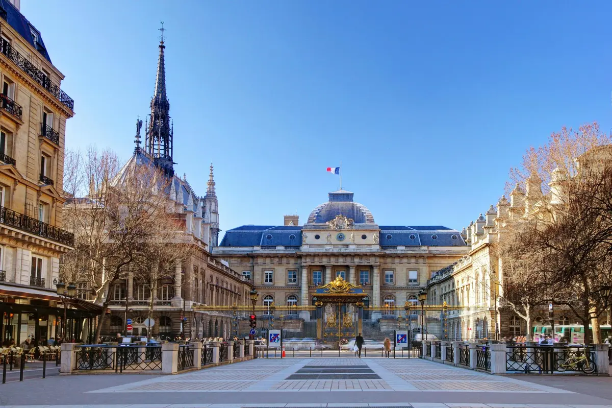 Bâtiment historique avec des portes ornées, le drapeau français au sommet, et une haute flèche à gauche, sous un ciel bleu clair.