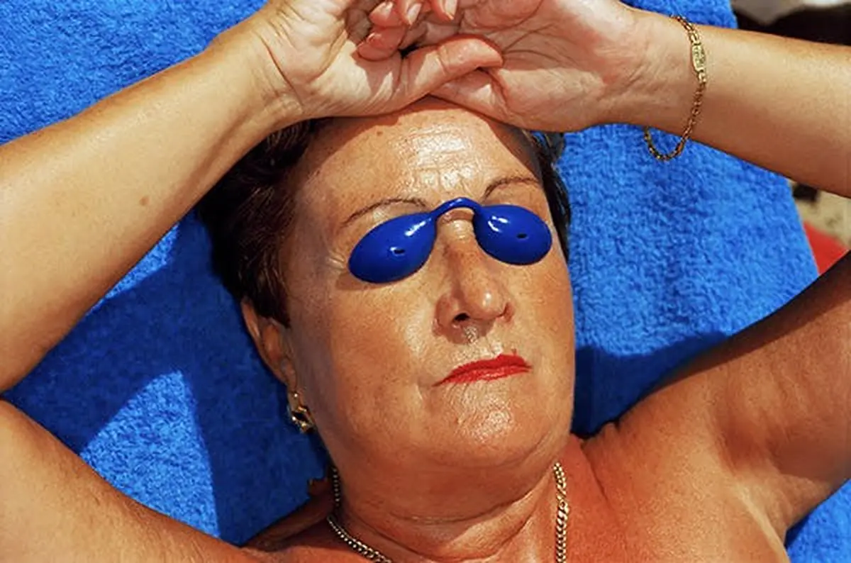 Une personne allongée, portant des lunettes de bronzage bleues, un collier et un bracelet, sur fond bleu.