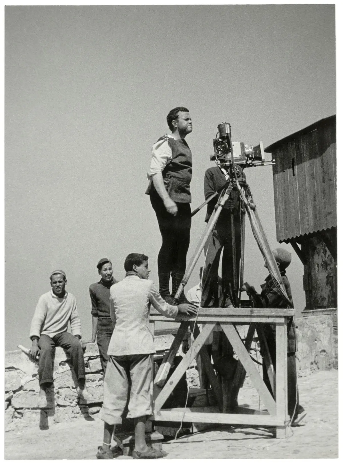 Un homme se tient sur une plate-forme en bois et manipule une caméra, tandis que plusieurs autres observent ou aident à proximité.