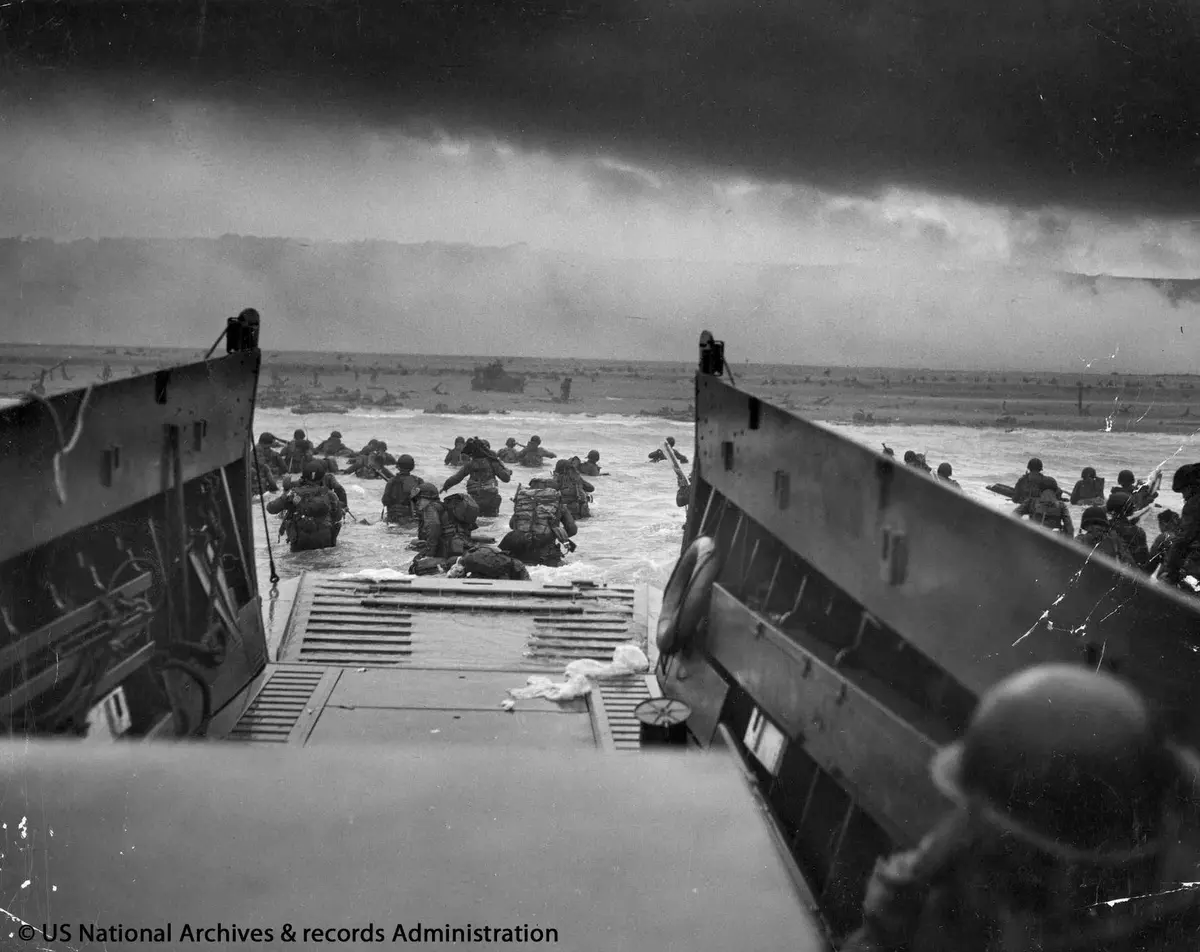 Soldats débarquant d'une péniche de débarquement dans l'eau, en direction d'une plage, sous un ciel nuageux.