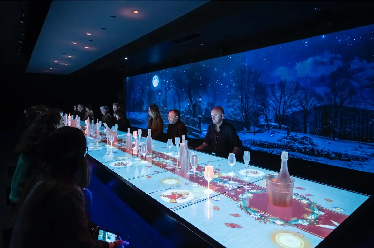 Des personnes assises à une longue table avec des projections lumineuses de nourriture et de boissons, sous une scène de ciel nocturne bleu foncé affichée au mur.