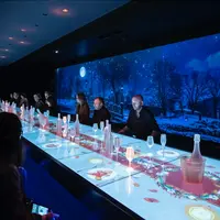 Des personnes assises à une longue table avec des projections lumineuses de nourriture et de boissons, sous une scène de ciel nocturne bleu foncé affichée au mur. &copy; ®Photo Anaka - Agence Clémence Farrell_Agence Sim&Sam