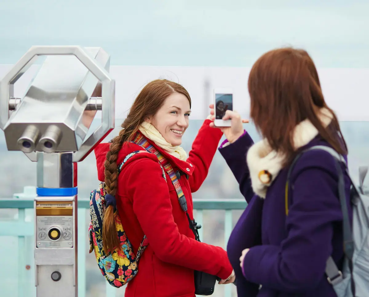 Deux femmes sourient, l'une prenant une photo de l'autre. Une jumelle à pièces est visible sur la gauche.
