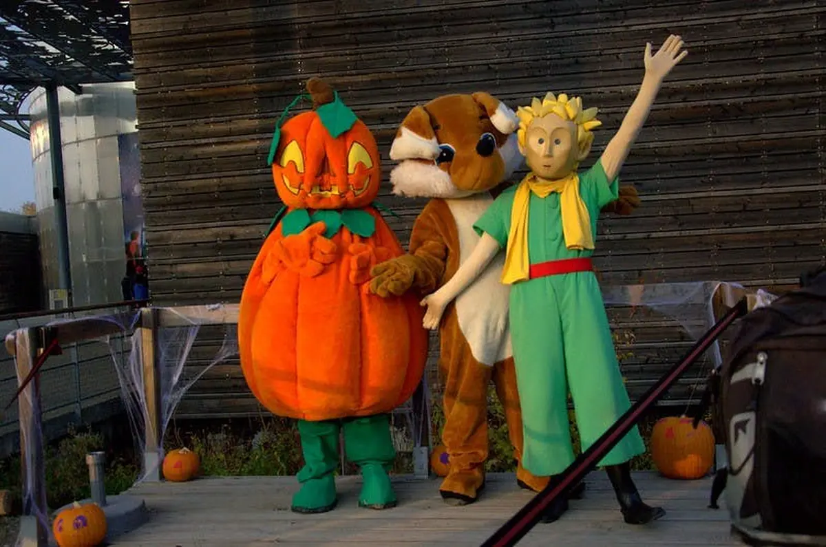 Trois personnes costumées se tiennent sur une scène : une citrouille, un écureuil et un personnage en costume vert avec une écharpe jaune.