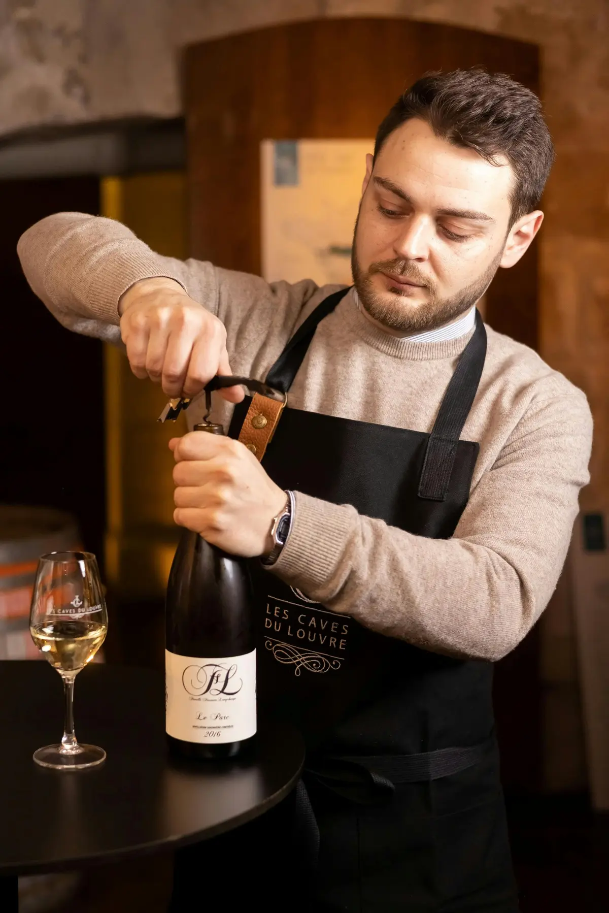 Un homme portant un pull et un tablier ouvre une bouteille de vin à l'aide d'un tire-bouchon dans un décor chaleureusement éclairé.