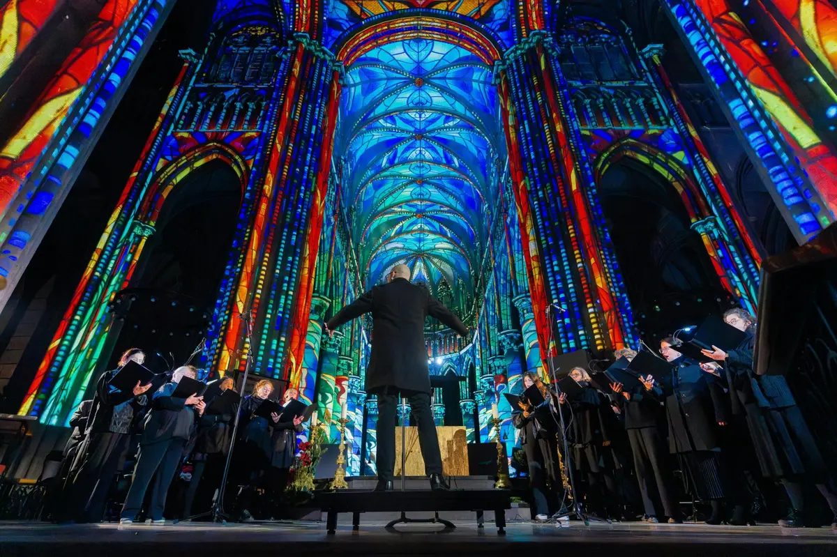 Un chef de chœur dirige une chorale à l'intérieur d'une cathédrale, dont les murs et le plafond sont ornés de projections de vitraux aux couleurs vives et éclatantes.