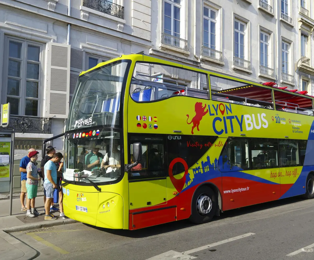 Un bus urbain lyonnais aux couleurs vives avec des passagers à bord, garé à côté d'un bâtiment par une journée ensoleillée.