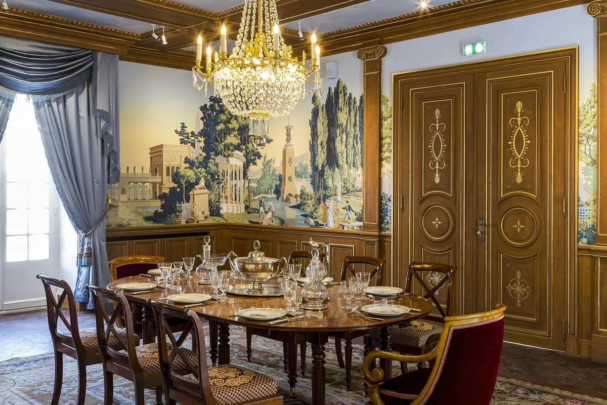 Élégante salle à manger avec une table en bois ornée de vaisselle et de cristaux, un lustre, des murs muraux et des portes en bois ornées.