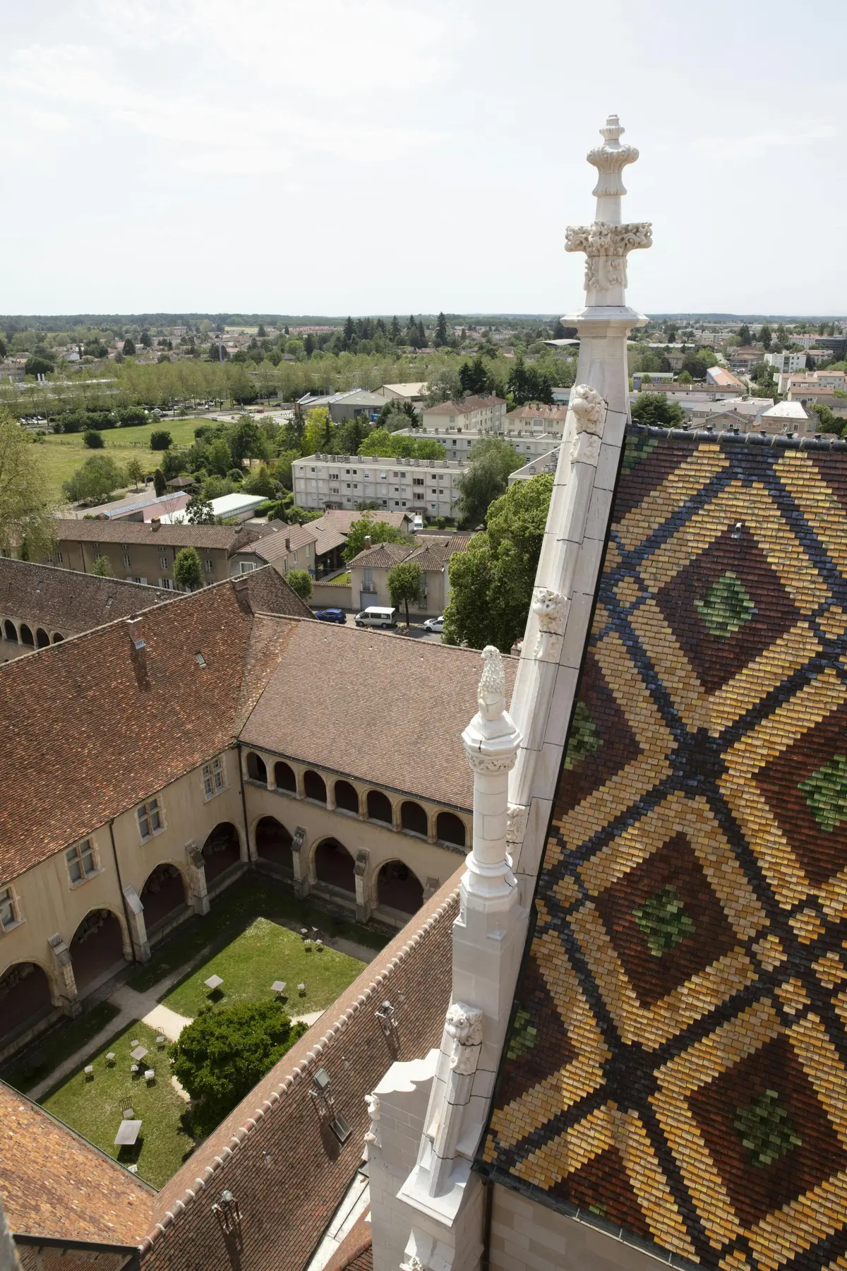 Toit en tuiles colorées d'un bâtiment historique, donnant sur une cour paisible avec des arches, et une ville avec de la verdure en arrière-plan.