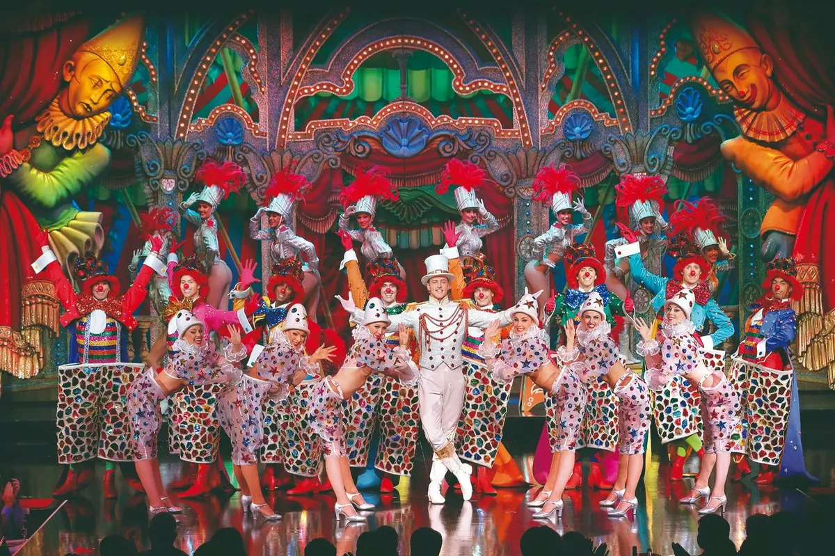 Un spectacle haut en couleur mettant en scène des artistes vêtus de costumes aux couleurs vives, avec en vedette un personnage vêtu d'un costume blanc et d'un haut-de-forme.