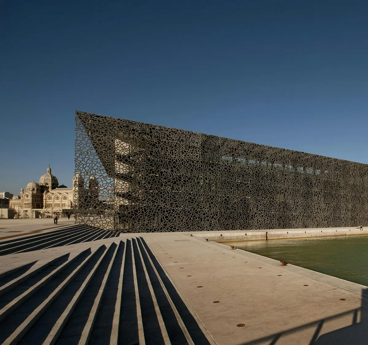 Bâtiment moderne avec un treillis complexe près d'un plan d'eau. Des marches et quelques personnes se trouvent au premier plan, avec un autre bâtiment historique à l'arrière-plan.