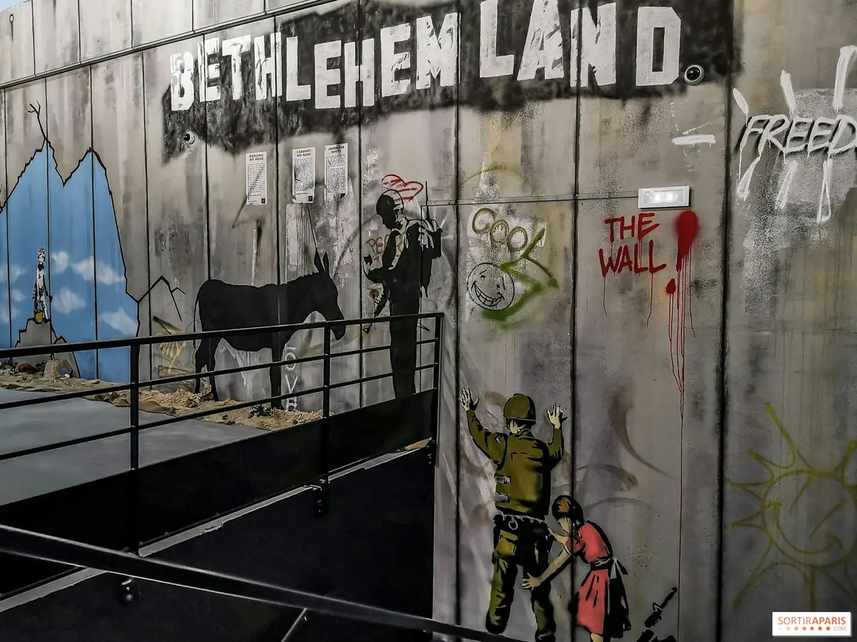 Un graffiti sur un mur montre des soldats, un âne et des enfants. Le texte indique BETHLEHEM LAND et THE WALL en grosses lettres.