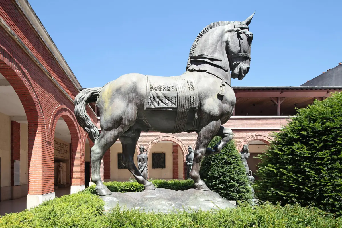 Grande statue de cheval dans une cour avec une colonnade de briques cintrées, des buissons et un ciel bleu clair.