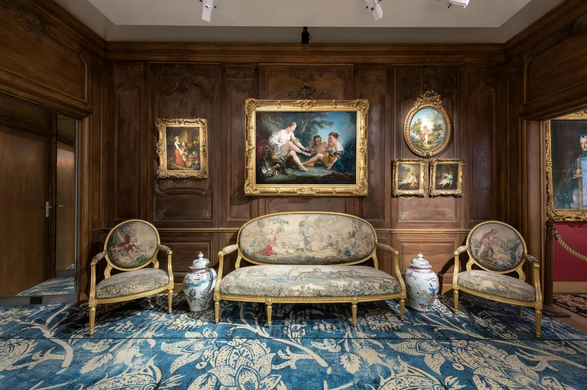 Salon orné de meubles anciens, de tableaux encadrés et de vases décoratifs posés sur un tapis bleu à motifs contre des murs lambrissés.