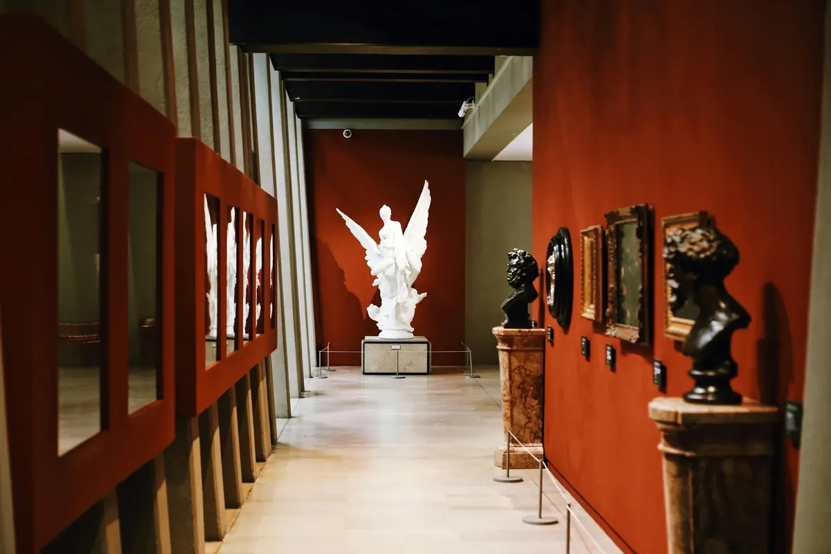 Un couloir de musée avec une statue ailée blanche au bout, des murs rouges, des miroirs à gauche, et diverses sculptures et œuvres d'art encadrées à droite.