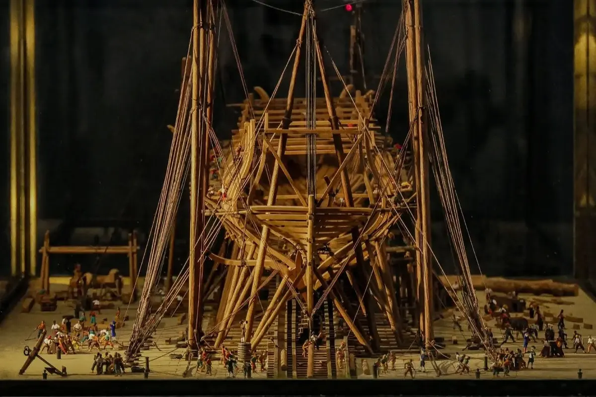 Un navire en bois détaillé en construction avec des personnages miniatures travaillant autour de lui, entouré d'échafaudages et de gréements.