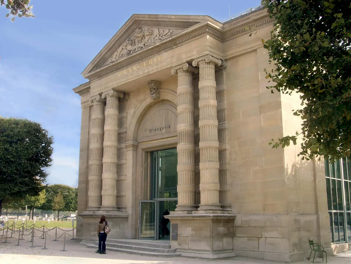 Une personne se tient devant un bâtiment néoclassique avec de hautes colonnes cannelées et une grande entrée voûtée portant l'inscription Orangerie.