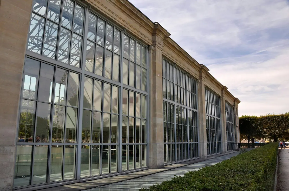 Un bâtiment à façade vitrée avec une armature métallique et de grandes fenêtres, sous un ciel nuageux, à côté d'une haie bien taillée.
