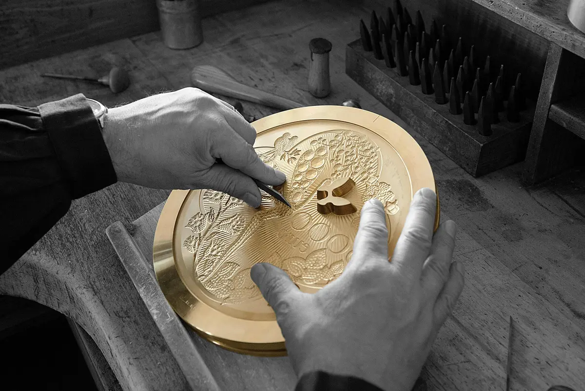 Des mains gravent un dessin détaillé sur une plaque circulaire en laiton sur un établi en bois avec plusieurs outils de gravure à proximité.