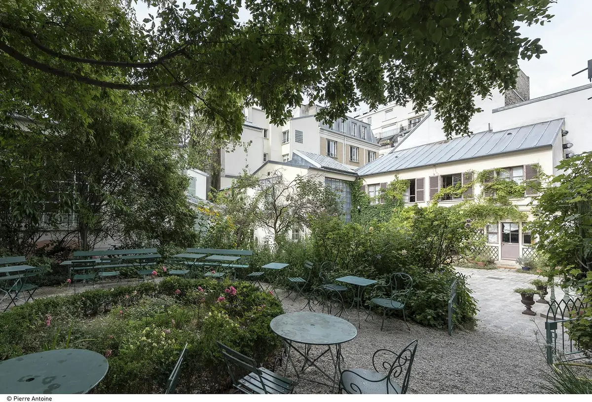 Un jardin extérieur avec des tables et des chaises en métal, entouré de plantes et d'arbres, avec des bâtiments blancs en arrière-plan.