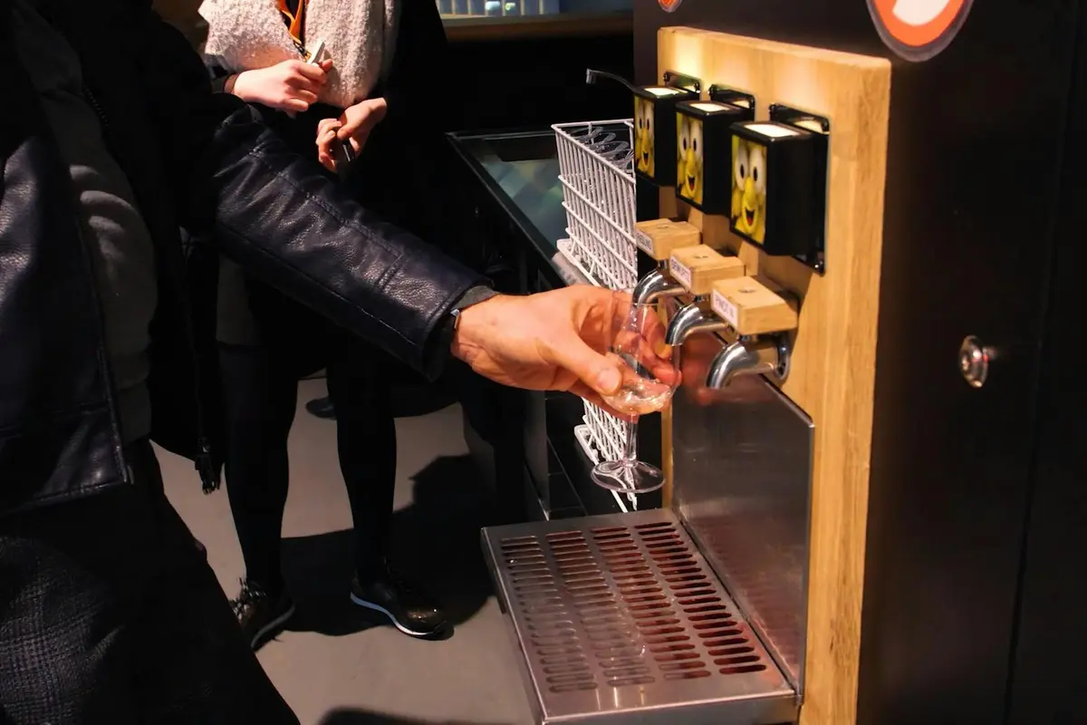 Une personne vêtue d'une veste en cuir remplit un verre à partir d'un robinet de bière dont les poignées sont ornées de visages de dessins animés dans un lieu public.