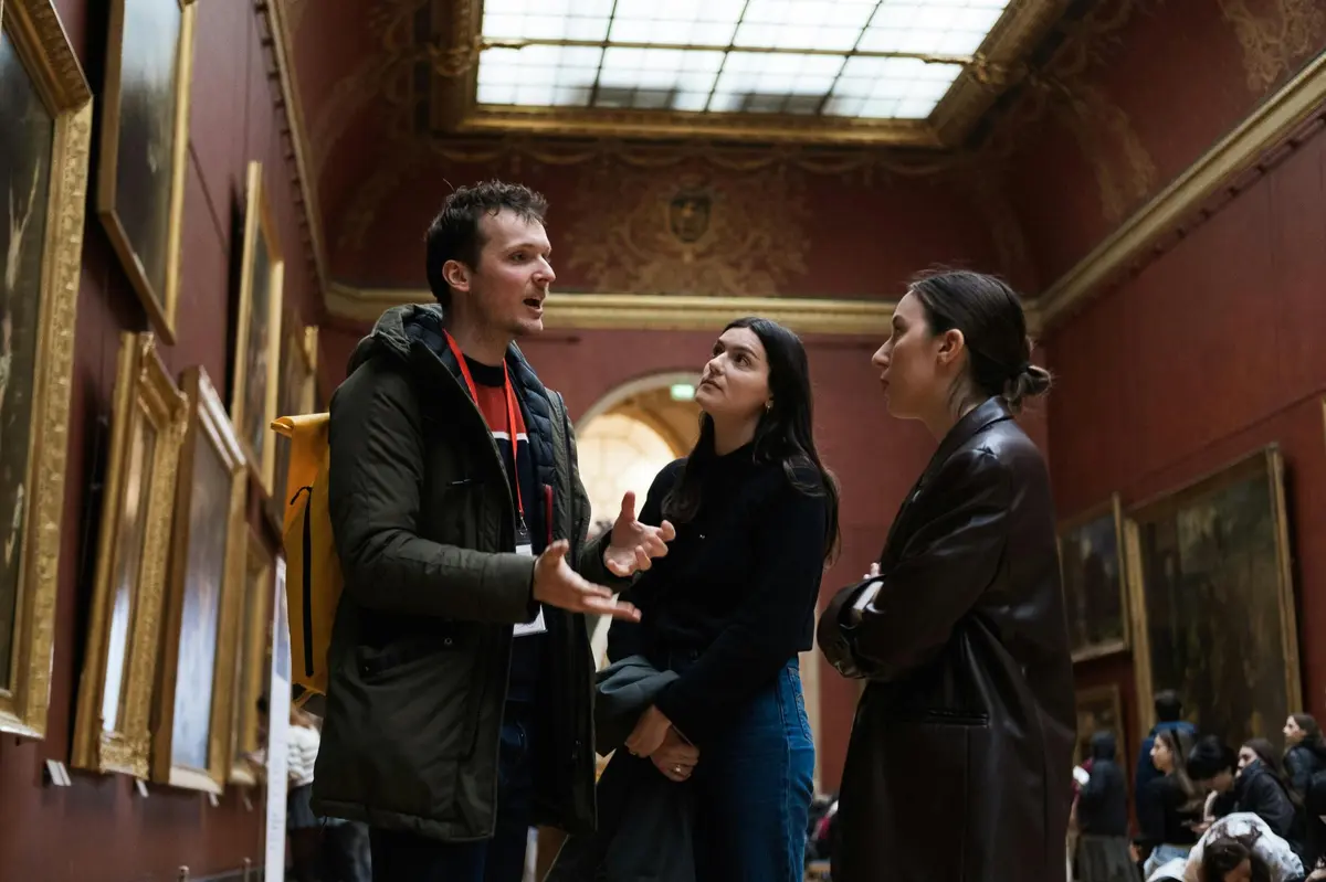 Three people are engaged in a conversation in a gallery with red walls, ornate decorations, and a large skylight.