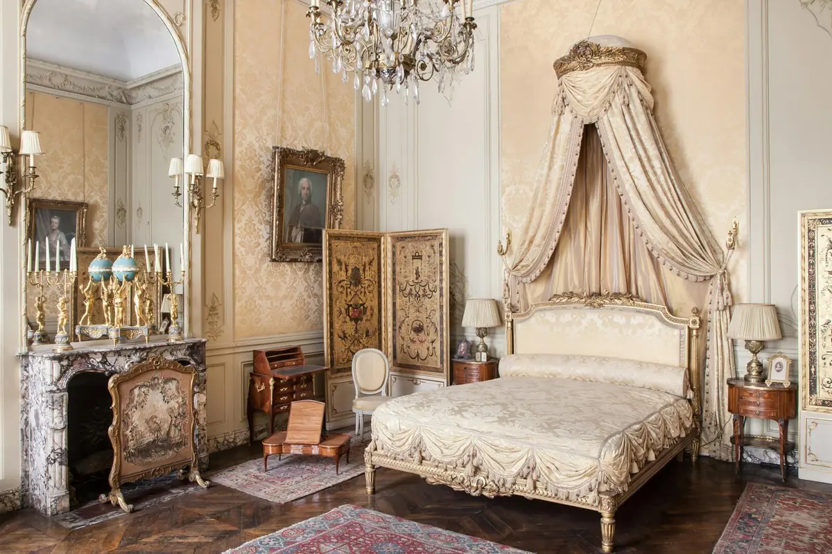 Chambre à coucher ornée d'un lustre, d'un lit à baldaquin, de meubles anciens, d'un grand miroir, d'un tapis et d'un portrait au mur.