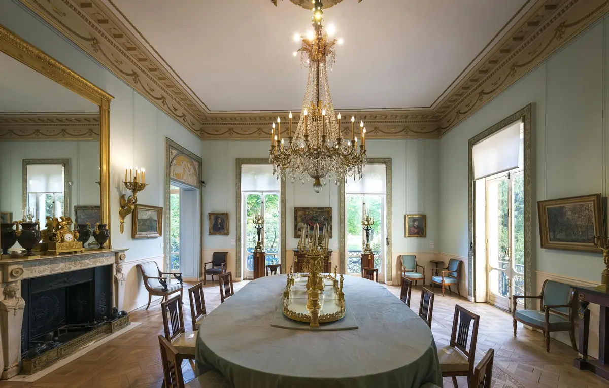 Salle à manger élégante avec un grand lustre, une table ovale avec un centre de table décoratif, entourée de chaises et de peintures sur les murs.